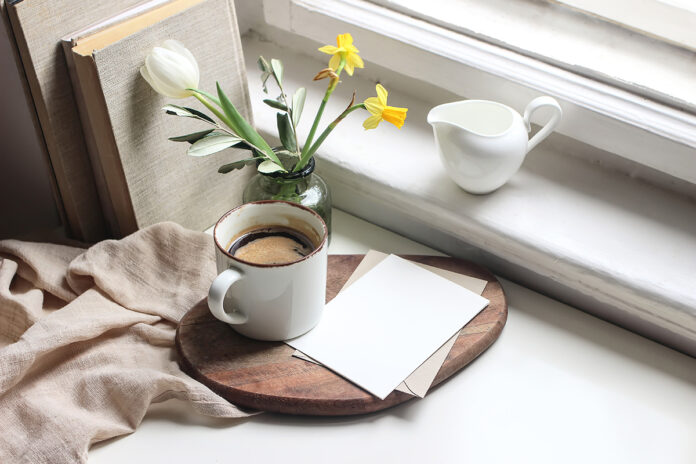 Cozy Easter spring still life. Greeting card mockup scene. Cup of coffee, books, wooden cutting board, milk pitcher and vase of flowers on windowsill. Floral composition. Yellow daffodils and tulip.
